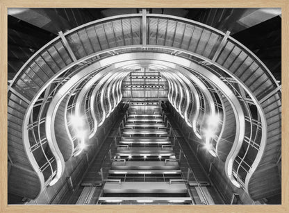 A symmetrical, low-angle black and white photograph of a futuristic escalator or grand staircase. The structure features illuminated, repeating curved arches on both sides, creating a tunnel-like perspective leading upwards. The image is presented within a silver-colored frame. Artwork