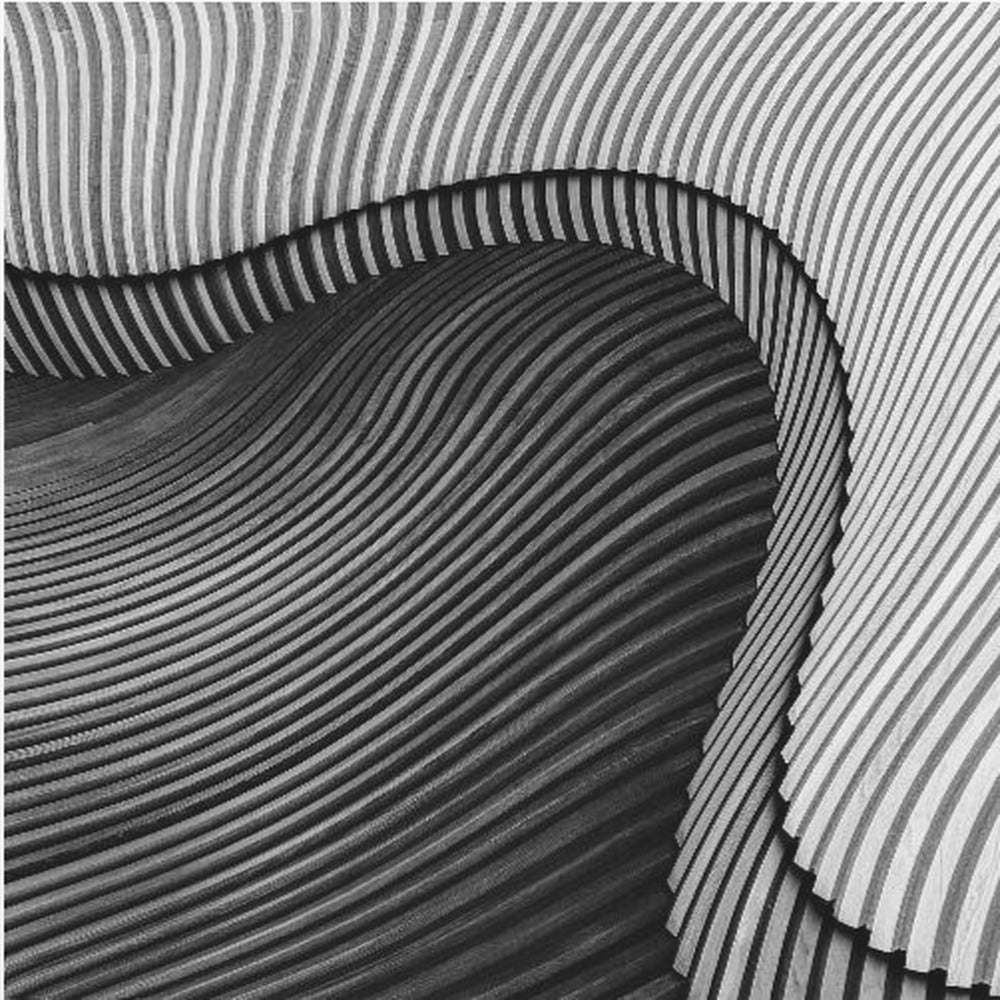 A square, black and white abstract photograph in a light wood frame. The image features layers of flowing, curved surfaces with parallel grooves, creating a strong sense of texture and depth. Lighter, striped layers with vertical lines curve over a darker, undulating section, resembling waves or sculpted wood, drawing the eye into a central dark crevice. Decor