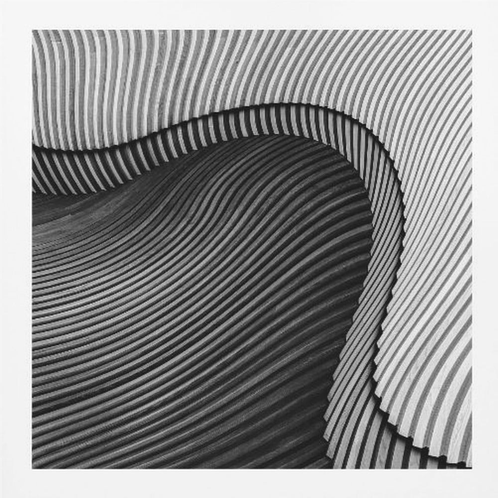 A square, black and white abstract photograph in a light wood frame. The image features layers of flowing, curved surfaces with parallel grooves, creating a strong sense of texture and depth. Lighter, striped layers with vertical lines curve over a darker, undulating section, resembling waves or sculpted wood, drawing the eye into a central dark crevice. Decor