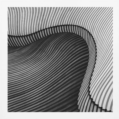 A square, black and white abstract photograph in a light wood frame. The image features layers of flowing, curved surfaces with parallel grooves, creating a strong sense of texture and depth. Lighter, striped layers with vertical lines curve over a darker, undulating section, resembling waves or sculpted wood, drawing the eye into a central dark crevice. Decor