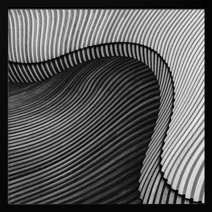 A square, black and white abstract photograph in a light wood frame. The image features layers of flowing, curved surfaces with parallel grooves, creating a strong sense of texture and depth. Lighter, striped layers with vertical lines curve over a darker, undulating section, resembling waves or sculpted wood, drawing the eye into a central dark crevice. Decor