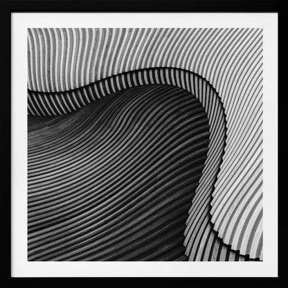 A square, black and white abstract photograph in a light wood frame. The image features layers of flowing, curved surfaces with parallel grooves, creating a strong sense of texture and depth. Lighter, striped layers with vertical lines curve over a darker, undulating section, resembling waves or sculpted wood, drawing the eye into a central dark crevice. Decor