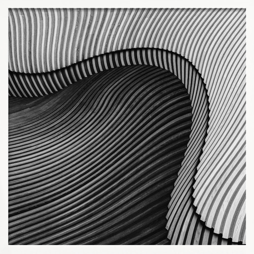 A square, black and white abstract photograph in a light wood frame. The image features layers of flowing, curved surfaces with parallel grooves, creating a strong sense of texture and depth. Lighter, striped layers with vertical lines curve over a darker, undulating section, resembling waves or sculpted wood, drawing the eye into a central dark crevice. Decor