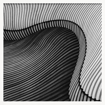 A square, black and white abstract photograph in a light wood frame. The image features layers of flowing, curved surfaces with parallel grooves, creating a strong sense of texture and depth. Lighter, striped layers with vertical lines curve over a darker, undulating section, resembling waves or sculpted wood, drawing the eye into a central dark crevice. Decor