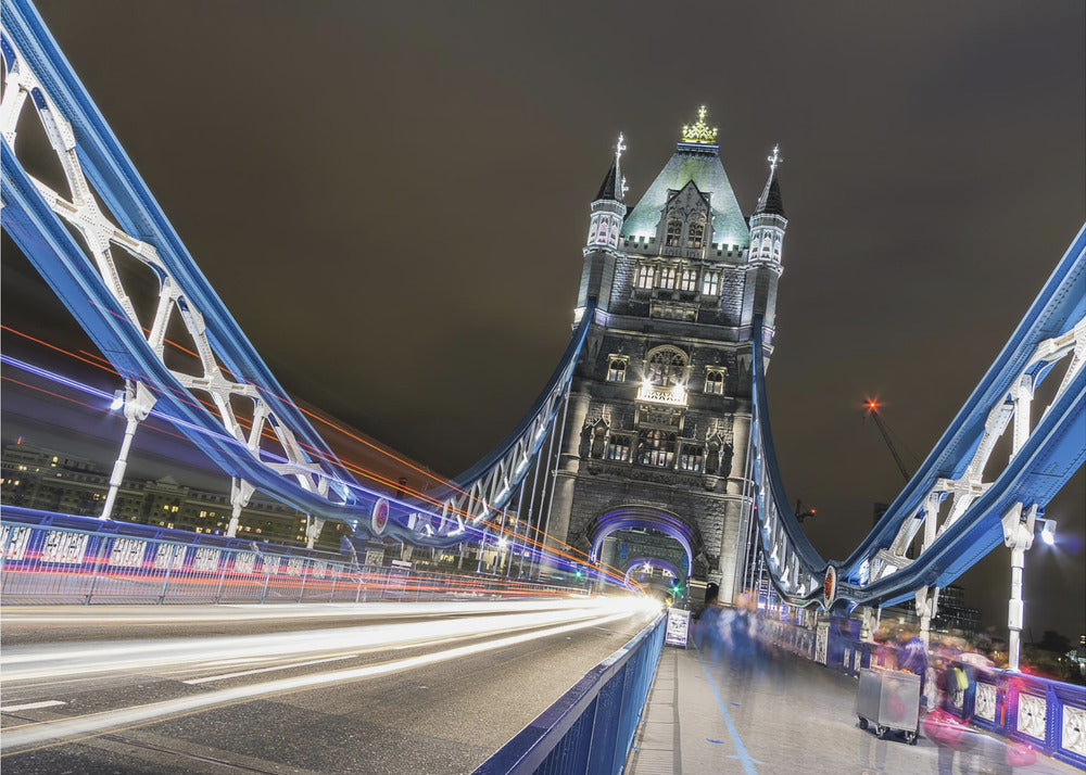 A dramatic, long-exposure night photograph from the deck of London's Tower Bridge, showing vibrant blue support structures curving towards an illuminated stone tower against a dark sky, with bright streaks of light from traffic. Wall Art