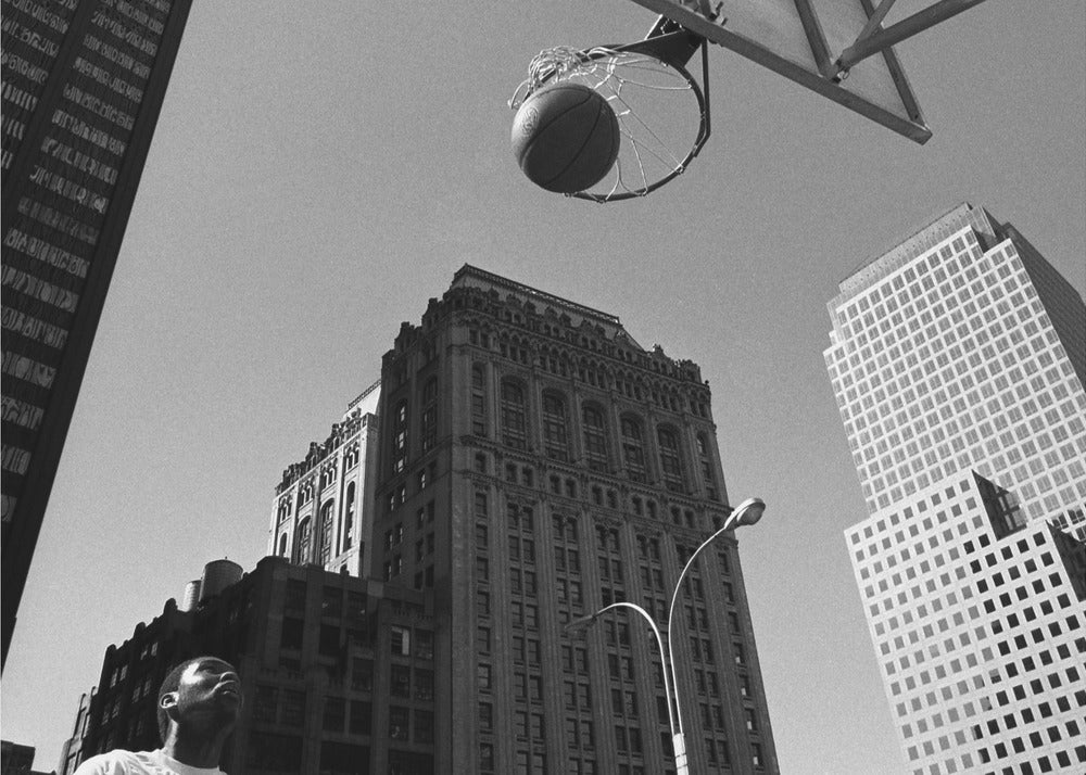 A low-angle, black and white photograph capturing a basketball as it's about to fall through the hoop, set against a backdrop of towering city skyscrapers. A player's face is visible in the bottom left corner, looking up at the ball. The entire image is presented within a silver-colored frame. Artwork