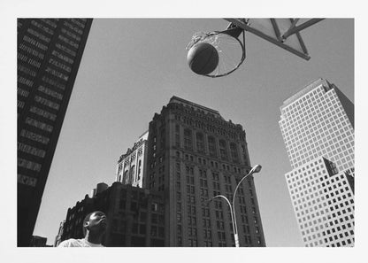 A low-angle, black and white photograph capturing a basketball as it's about to fall through the hoop, set against a backdrop of towering city skyscrapers. A player's face is visible in the bottom left corner, looking up at the ball. The entire image is presented within a silver-colored frame. Artwork