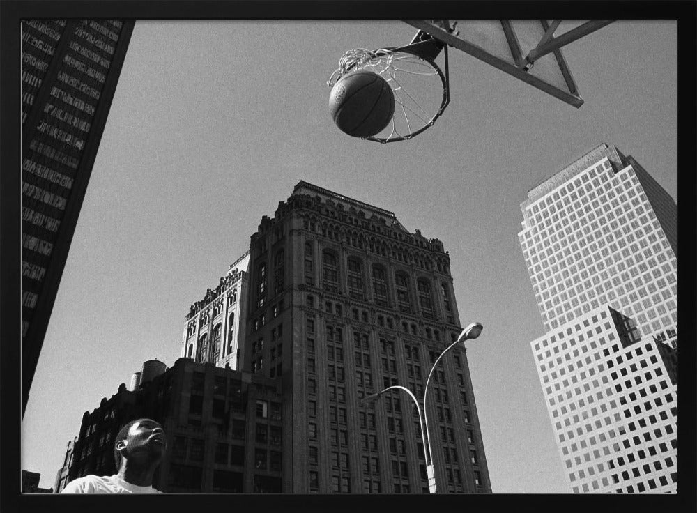 A low-angle, black and white photograph capturing a basketball as it's about to fall through the hoop, set against a backdrop of towering city skyscrapers. A player's face is visible in the bottom left corner, looking up at the ball. The entire image is presented within a silver-colored frame. Artwork