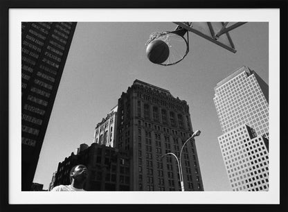 A low-angle, black and white photograph capturing a basketball as it's about to fall through the hoop, set against a backdrop of towering city skyscrapers. A player's face is visible in the bottom left corner, looking up at the ball. The entire image is presented within a silver-colored frame. Artwork