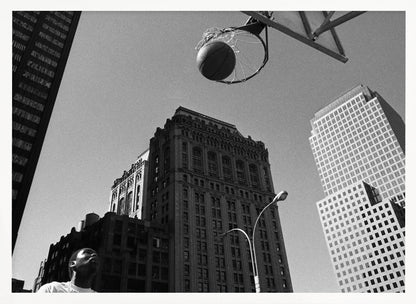 A low-angle, black and white photograph capturing a basketball as it's about to fall through the hoop, set against a backdrop of towering city skyscrapers. A player's face is visible in the bottom left corner, looking up at the ball. The entire image is presented within a silver-colored frame. Artwork
