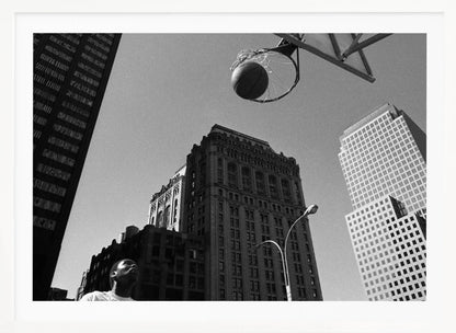 A low-angle, black and white photograph capturing a basketball as it's about to fall through the hoop, set against a backdrop of towering city skyscrapers. A player's face is visible in the bottom left corner, looking up at the ball. The entire image is presented within a silver-colored frame. Artwork