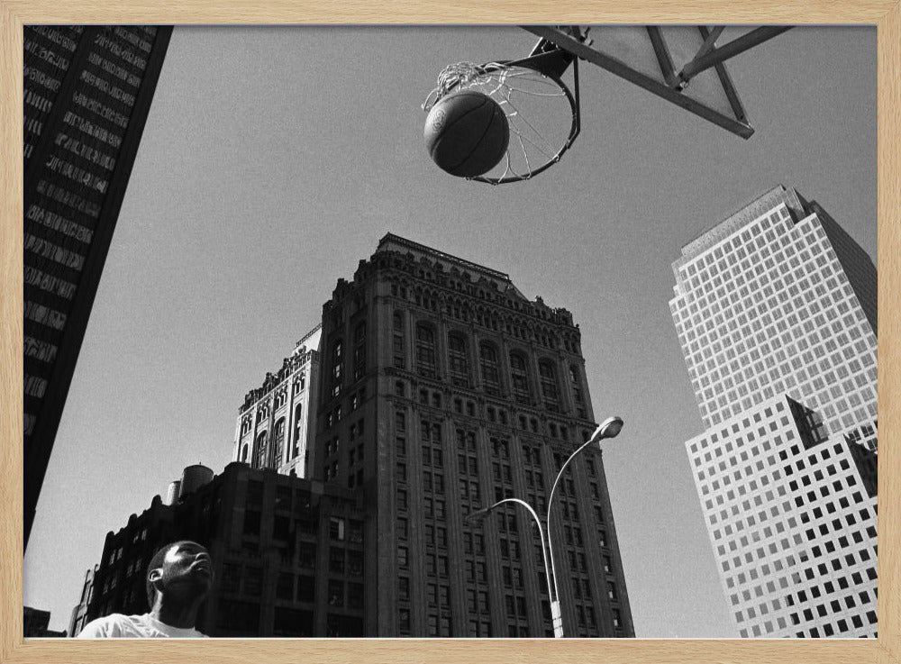 A low-angle, black and white photograph capturing a basketball as it's about to fall through the hoop, set against a backdrop of towering city skyscrapers. A player's face is visible in the bottom left corner, looking up at the ball. The entire image is presented within a silver-colored frame. Artwork
