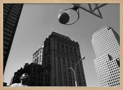 A low-angle, black and white photograph capturing a basketball as it's about to fall through the hoop, set against a backdrop of towering city skyscrapers. A player's face is visible in the bottom left corner, looking up at the ball. The entire image is presented within a silver-colored frame. Artwork