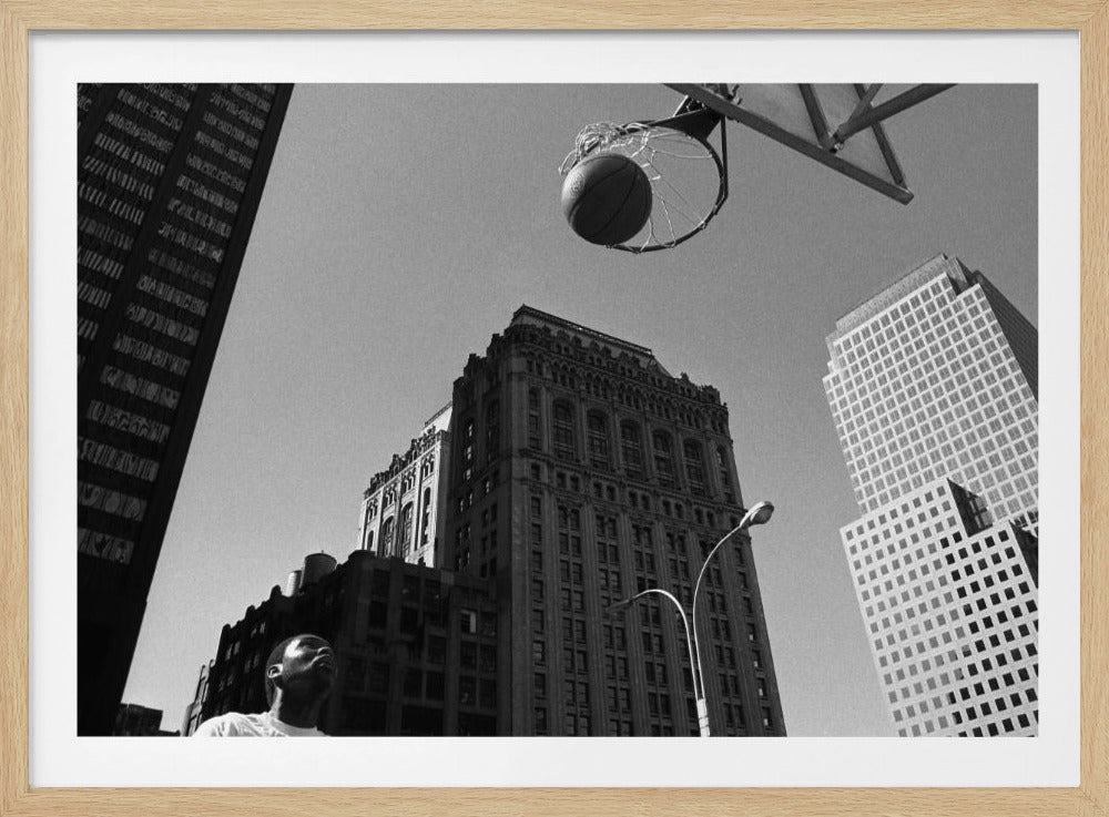 A low-angle, black and white photograph capturing a basketball as it's about to fall through the hoop, set against a backdrop of towering city skyscrapers. A player's face is visible in the bottom left corner, looking up at the ball. The entire image is presented within a silver-colored frame. Artwork