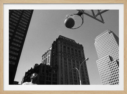 A low-angle, black and white photograph capturing a basketball as it's about to fall through the hoop, set against a backdrop of towering city skyscrapers. A player's face is visible in the bottom left corner, looking up at the ball. The entire image is presented within a silver-colored frame. Artwork