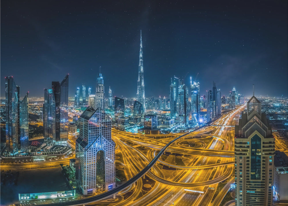 A vibrant aerial photograph of the Dubai skyline at night, with the iconic Burj Khalifa towering in the center. A complex web of brightly lit highways with light trails from traffic dominates the foreground, weaving between modern skyscrapers under a starry night sky. Decor