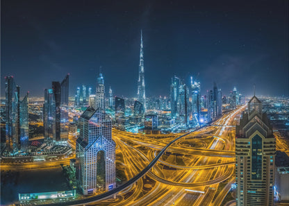A vibrant aerial photograph of the Dubai skyline at night, with the iconic Burj Khalifa towering in the center. A complex web of brightly lit highways with light trails from traffic dominates the foreground, weaving between modern skyscrapers under a starry night sky. Decor