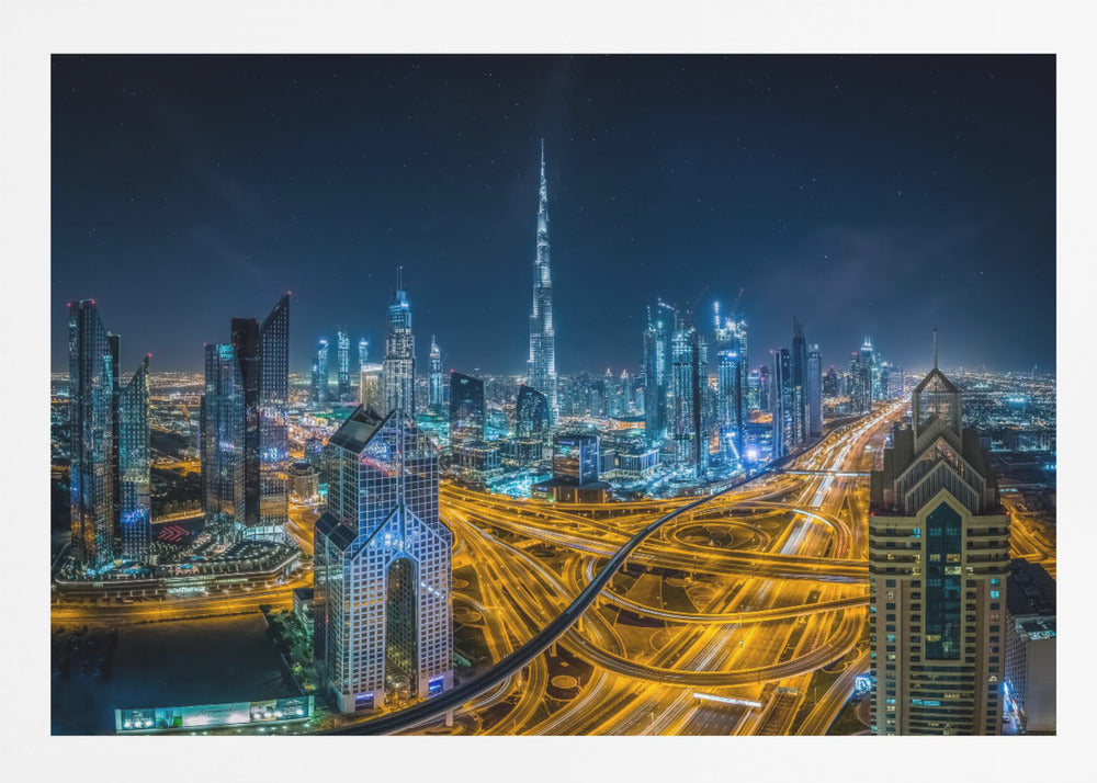 A vibrant aerial photograph of the Dubai skyline at night, with the iconic Burj Khalifa towering in the center. A complex web of brightly lit highways with light trails from traffic dominates the foreground, weaving between modern skyscrapers under a starry night sky. Decor