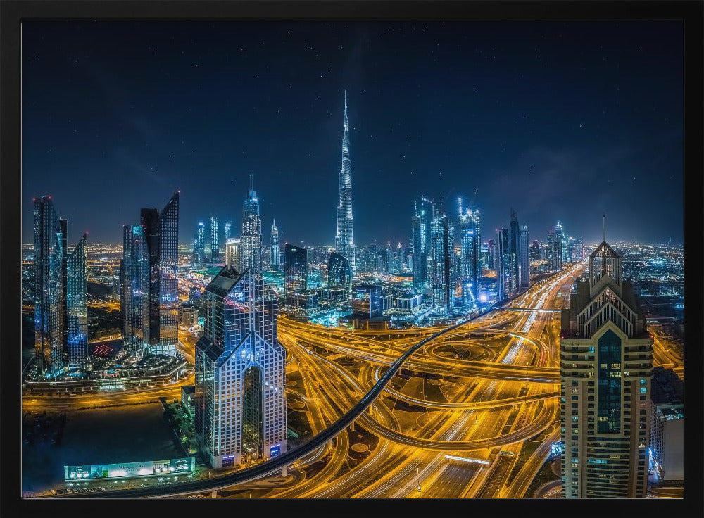 A vibrant aerial photograph of the Dubai skyline at night, with the iconic Burj Khalifa towering in the center. A complex web of brightly lit highways with light trails from traffic dominates the foreground, weaving between modern skyscrapers under a starry night sky. Decor