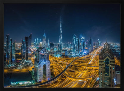 A vibrant aerial photograph of the Dubai skyline at night, with the iconic Burj Khalifa towering in the center. A complex web of brightly lit highways with light trails from traffic dominates the foreground, weaving between modern skyscrapers under a starry night sky. Decor