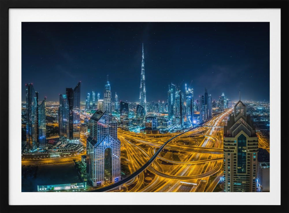 A vibrant aerial photograph of the Dubai skyline at night, with the iconic Burj Khalifa towering in the center. A complex web of brightly lit highways with light trails from traffic dominates the foreground, weaving between modern skyscrapers under a starry night sky. Decor