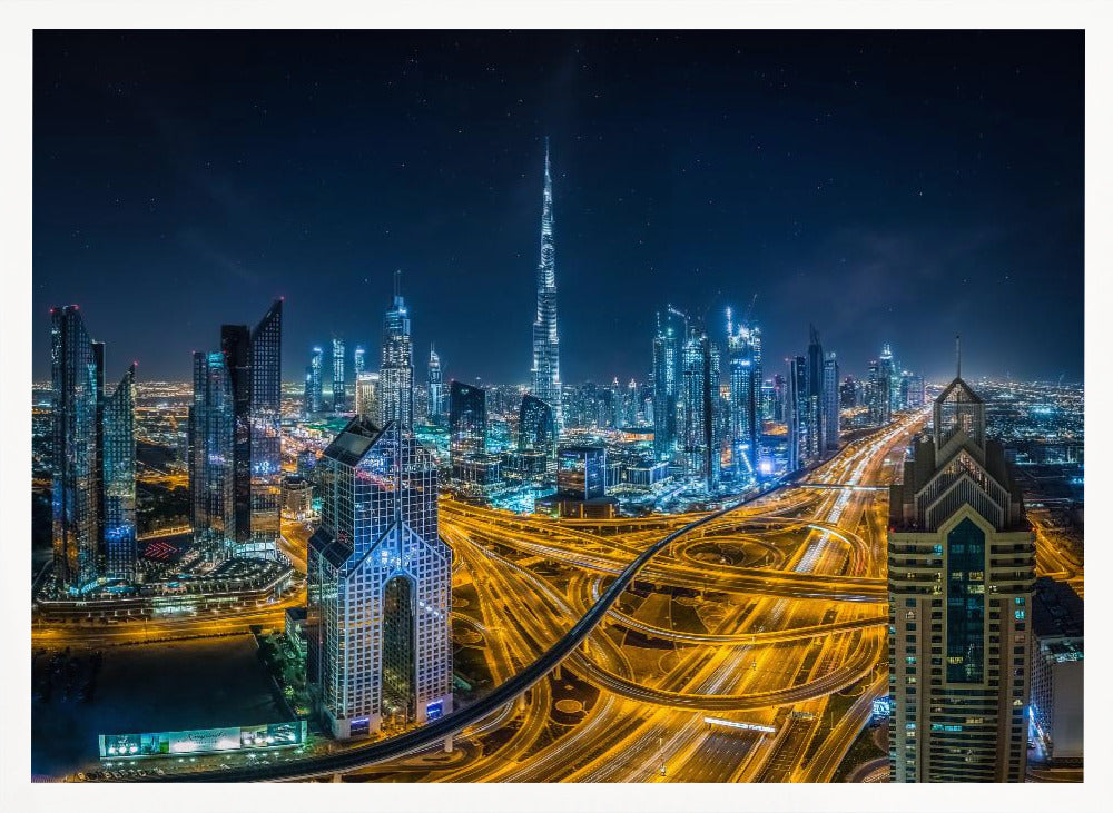 A vibrant aerial photograph of the Dubai skyline at night, with the iconic Burj Khalifa towering in the center. A complex web of brightly lit highways with light trails from traffic dominates the foreground, weaving between modern skyscrapers under a starry night sky. Decor