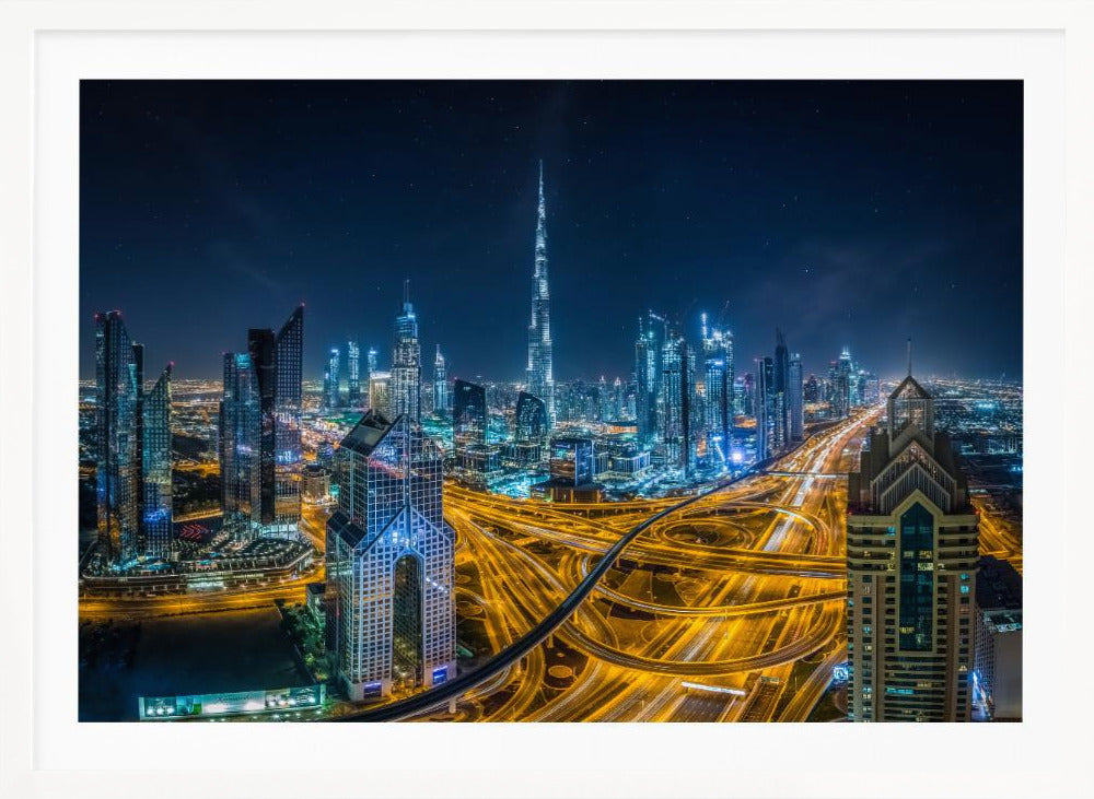 A vibrant aerial photograph of the Dubai skyline at night, with the iconic Burj Khalifa towering in the center. A complex web of brightly lit highways with light trails from traffic dominates the foreground, weaving between modern skyscrapers under a starry night sky. Decor