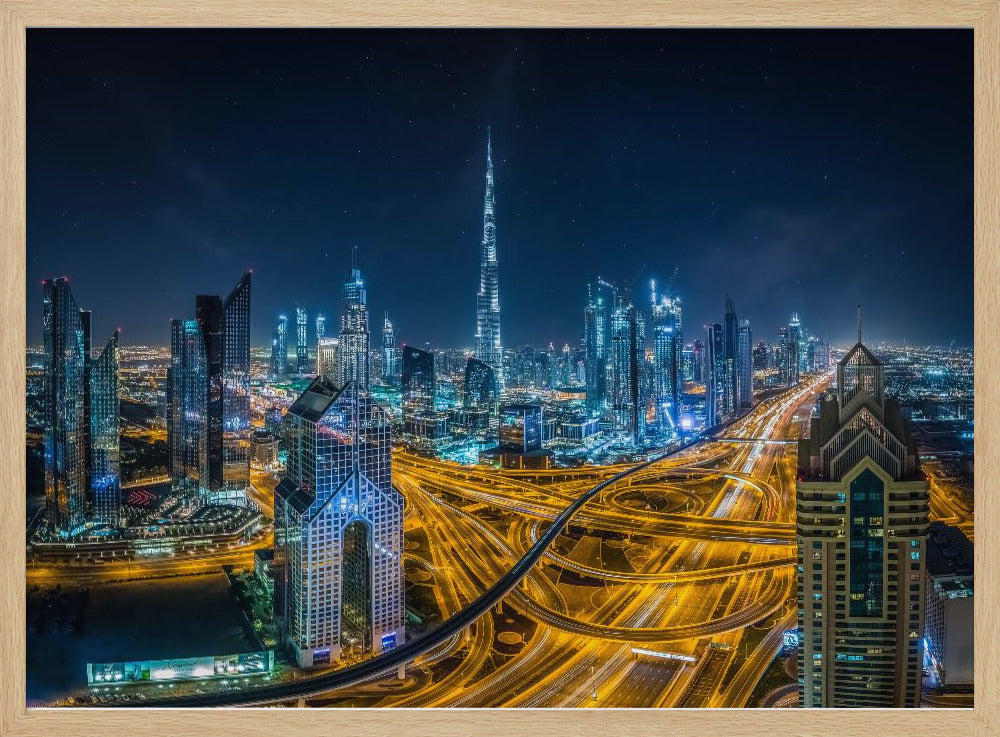 A vibrant aerial photograph of the Dubai skyline at night, with the iconic Burj Khalifa towering in the center. A complex web of brightly lit highways with light trails from traffic dominates the foreground, weaving between modern skyscrapers under a starry night sky. Decor