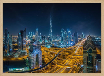 A vibrant aerial photograph of the Dubai skyline at night, with the iconic Burj Khalifa towering in the center. A complex web of brightly lit highways with light trails from traffic dominates the foreground, weaving between modern skyscrapers under a starry night sky. Decor
