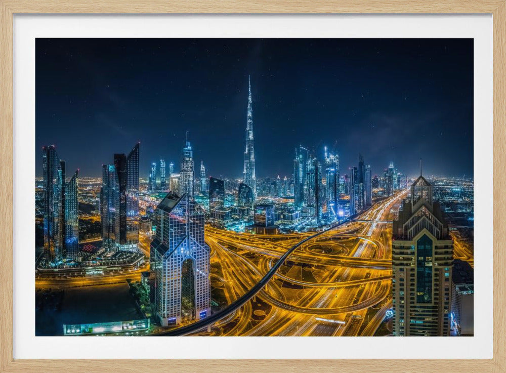 A vibrant aerial photograph of the Dubai skyline at night, with the iconic Burj Khalifa towering in the center. A complex web of brightly lit highways with light trails from traffic dominates the foreground, weaving between modern skyscrapers under a starry night sky. Decor