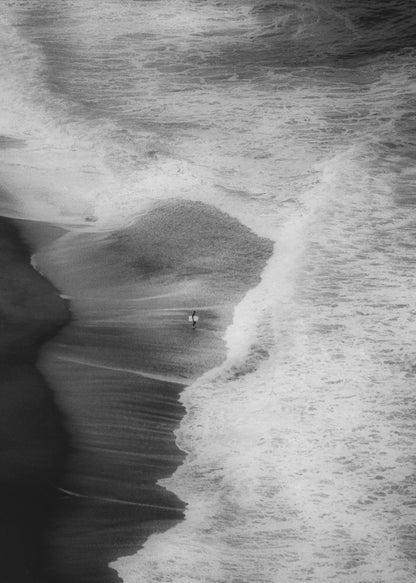 A high-angle, black and white photograph of a lone surfer carrying a surfboard at the water's edge on a dark sand beach. The vast, textured ocean with large, foamy waves crashing ashore dwarfs the solitary figure, creating a sense of scale and solitude. Decor