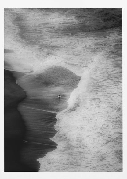 A high-angle, black and white photograph of a lone surfer carrying a surfboard at the water's edge on a dark sand beach. The vast, textured ocean with large, foamy waves crashing ashore dwarfs the solitary figure, creating a sense of scale and solitude. Decor
