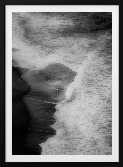 A high-angle, black and white photograph of a lone surfer carrying a surfboard at the water's edge on a dark sand beach. The vast, textured ocean with large, foamy waves crashing ashore dwarfs the solitary figure, creating a sense of scale and solitude. Decor