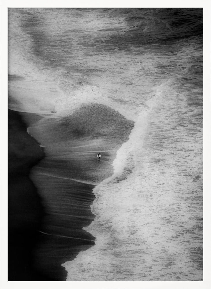 A high-angle, black and white photograph of a lone surfer carrying a surfboard at the water's edge on a dark sand beach. The vast, textured ocean with large, foamy waves crashing ashore dwarfs the solitary figure, creating a sense of scale and solitude. Decor