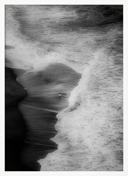 A high-angle, black and white photograph of a lone surfer carrying a surfboard at the water's edge on a dark sand beach. The vast, textured ocean with large, foamy waves crashing ashore dwarfs the solitary figure, creating a sense of scale and solitude. Decor
