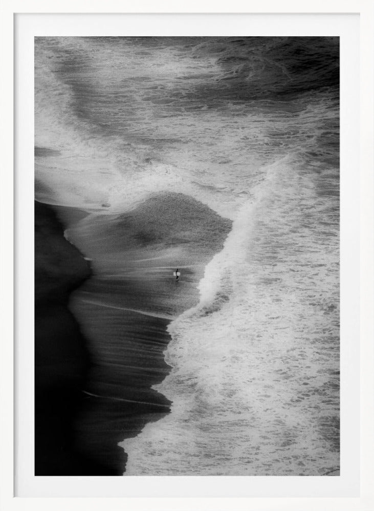 A high-angle, black and white photograph of a lone surfer carrying a surfboard at the water's edge on a dark sand beach. The vast, textured ocean with large, foamy waves crashing ashore dwarfs the solitary figure, creating a sense of scale and solitude. Decor