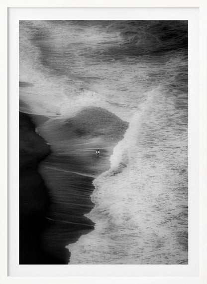 A high-angle, black and white photograph of a lone surfer carrying a surfboard at the water's edge on a dark sand beach. The vast, textured ocean with large, foamy waves crashing ashore dwarfs the solitary figure, creating a sense of scale and solitude. Decor