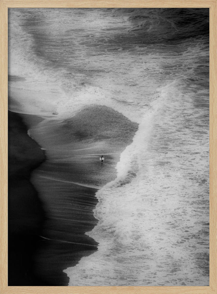 A high-angle, black and white photograph of a lone surfer carrying a surfboard at the water's edge on a dark sand beach. The vast, textured ocean with large, foamy waves crashing ashore dwarfs the solitary figure, creating a sense of scale and solitude. Decor