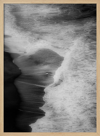 A high-angle, black and white photograph of a lone surfer carrying a surfboard at the water's edge on a dark sand beach. The vast, textured ocean with large, foamy waves crashing ashore dwarfs the solitary figure, creating a sense of scale and solitude. Decor