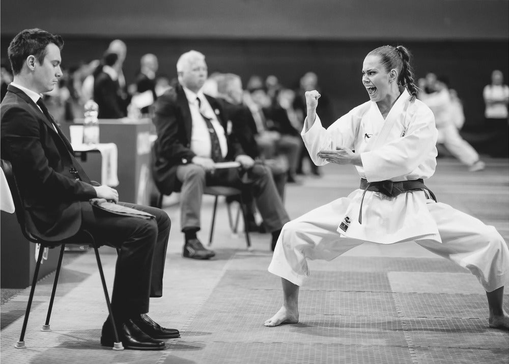 A dynamic black and white photograph of a female martial artist in a white gi and black belt, mid-kiai. She is in a deep, wide stance on a matted floor, her face filled with intensity and her fist clenched. In the foreground, a man in a suit sits and watches, while other spectators are seated in the blurred background, suggesting a competition or demonstration setting. The entire image has a silver-colored frame. Print