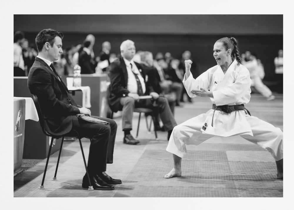 A dynamic black and white photograph of a female martial artist in a white gi and black belt, mid-kiai. She is in a deep, wide stance on a matted floor, her face filled with intensity and her fist clenched. In the foreground, a man in a suit sits and watches, while other spectators are seated in the blurred background, suggesting a competition or demonstration setting. The entire image has a silver-colored frame. Print