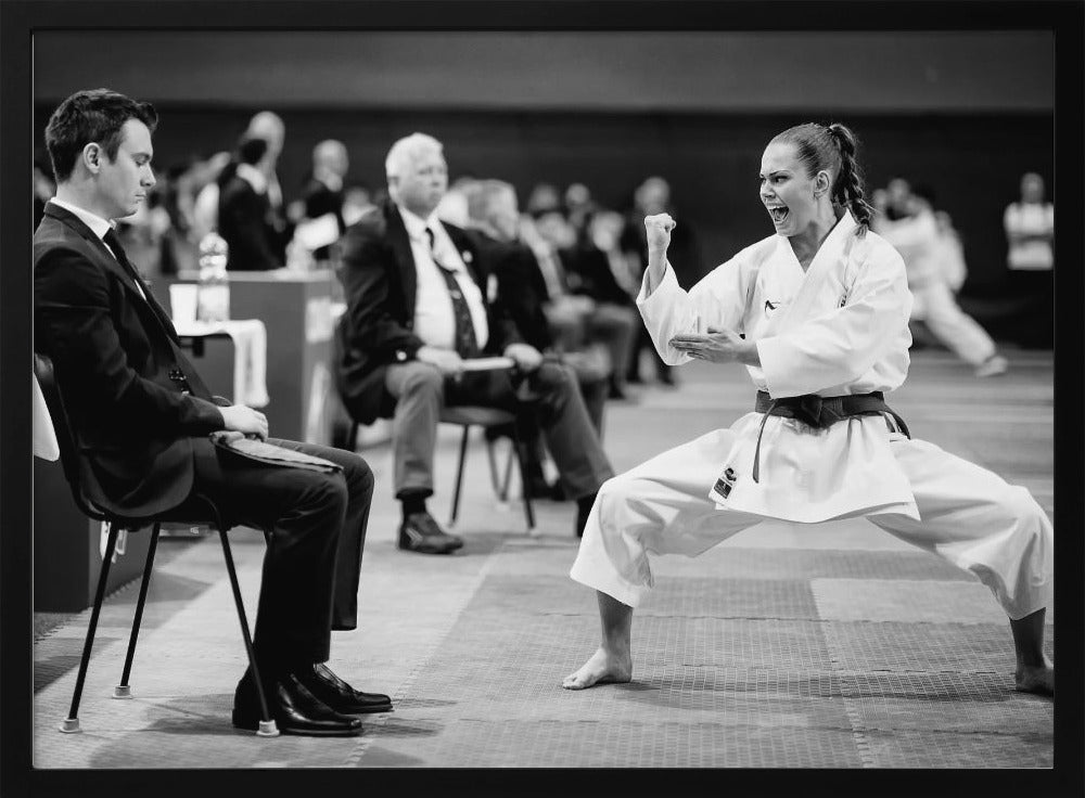 A dynamic black and white photograph of a female martial artist in a white gi and black belt, mid-kiai. She is in a deep, wide stance on a matted floor, her face filled with intensity and her fist clenched. In the foreground, a man in a suit sits and watches, while other spectators are seated in the blurred background, suggesting a competition or demonstration setting. The entire image has a silver-colored frame. Print