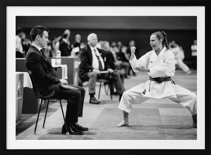 A dynamic black and white photograph of a female martial artist in a white gi and black belt, mid-kiai. She is in a deep, wide stance on a matted floor, her face filled with intensity and her fist clenched. In the foreground, a man in a suit sits and watches, while other spectators are seated in the blurred background, suggesting a competition or demonstration setting. The entire image has a silver-colored frame. Print