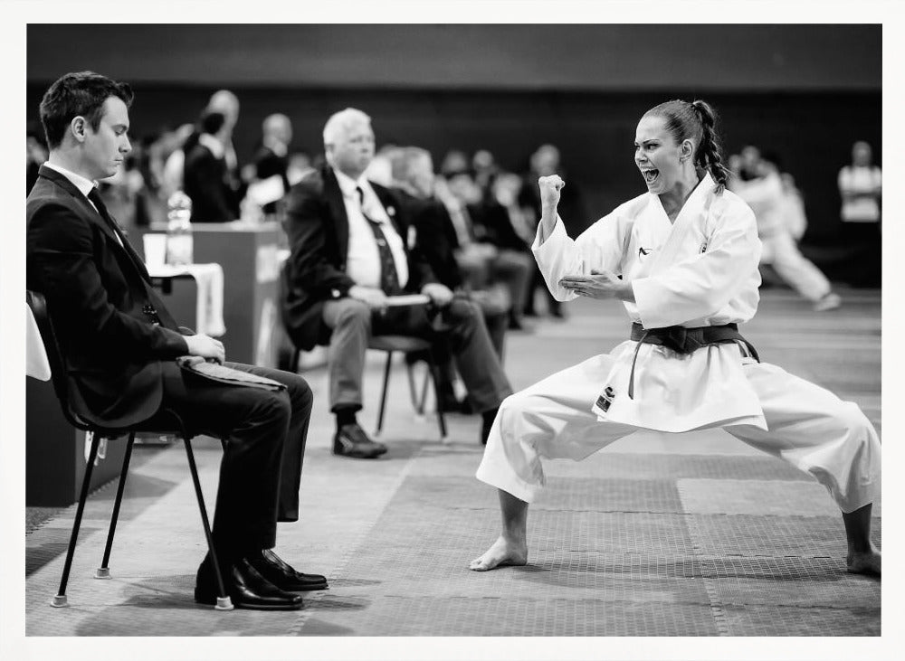 A dynamic black and white photograph of a female martial artist in a white gi and black belt, mid-kiai. She is in a deep, wide stance on a matted floor, her face filled with intensity and her fist clenched. In the foreground, a man in a suit sits and watches, while other spectators are seated in the blurred background, suggesting a competition or demonstration setting. The entire image has a silver-colored frame. Print