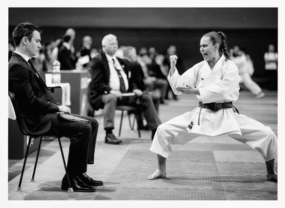 A dynamic black and white photograph of a female martial artist in a white gi and black belt, mid-kiai. She is in a deep, wide stance on a matted floor, her face filled with intensity and her fist clenched. In the foreground, a man in a suit sits and watches, while other spectators are seated in the blurred background, suggesting a competition or demonstration setting. The entire image has a silver-colored frame. Print