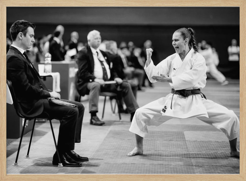A dynamic black and white photograph of a female martial artist in a white gi and black belt, mid-kiai. She is in a deep, wide stance on a matted floor, her face filled with intensity and her fist clenched. In the foreground, a man in a suit sits and watches, while other spectators are seated in the blurred background, suggesting a competition or demonstration setting. The entire image has a silver-colored frame. Print