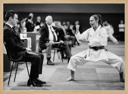 A dynamic black and white photograph of a female martial artist in a white gi and black belt, mid-kiai. She is in a deep, wide stance on a matted floor, her face filled with intensity and her fist clenched. In the foreground, a man in a suit sits and watches, while other spectators are seated in the blurred background, suggesting a competition or demonstration setting. The entire image has a silver-colored frame. Print