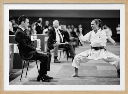 A dynamic black and white photograph of a female martial artist in a white gi and black belt, mid-kiai. She is in a deep, wide stance on a matted floor, her face filled with intensity and her fist clenched. In the foreground, a man in a suit sits and watches, while other spectators are seated in the blurred background, suggesting a competition or demonstration setting. The entire image has a silver-colored frame. Print