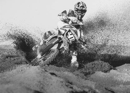 A dynamic black and white action photo of a motocross rider in full gear leaning hard into a turn, kicking up a large spray of dirt and sand from the track. The image is presented within a silver frame. Decor