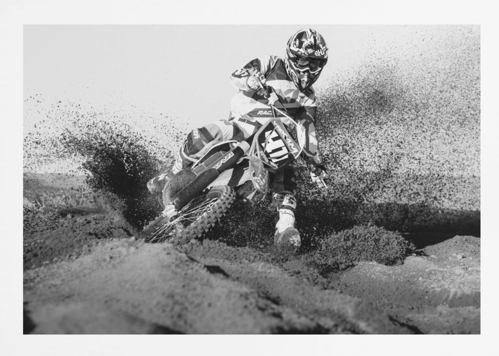A dynamic black and white action photo of a motocross rider in full gear leaning hard into a turn, kicking up a large spray of dirt and sand from the track. The image is presented within a silver frame. Decor
